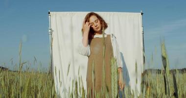 A young beautiful red-haired woman poses against a white screen set among a wheat field, looking into the camera and straightening her hair with her hands. 4k , red komodo video