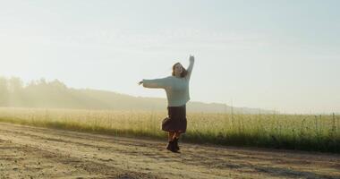 A beautiful young woman dressed in a warm sweater laughs and circles around on a gravel road near an agricultural field, enjoying the warm rays of the rising sun. 4k, , red komodo video