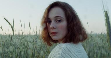 A young red-haired woman of Caucasian appearance turns her head and looks directly into the camera with a slight smile while standing among tall ears in a wheat field, close-up. 4k , red komodo video