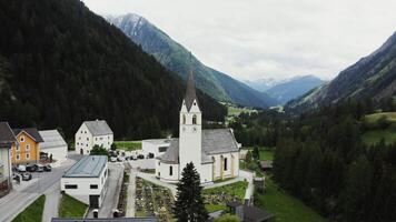 A small Catholic church with a parish cemetery in the courtyard, located in a lowland between the Alpine mountains with slopes overgrown with pine forests. Drone , Austria video