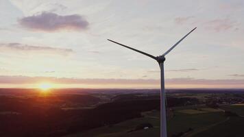 A close-up of the rotating blades of a wind turbine illuminated by the rays of the rising sun. Scenic green valley aerial view, drone video
