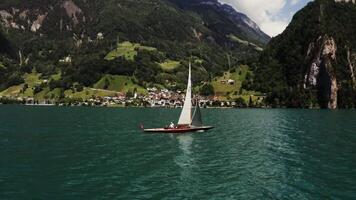 Scenic drone footage of a sailing boat with a Swiss flag sailing on a beautiful lake at the foot of the Alps, not far from a village located on the coast video