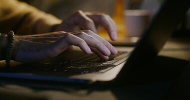 un' di carnagione chiara uomo vestito nel casuale Abiti è digitando su un' il computer portatile mentre seduta a un' tavolo di il leggero di il lampade. avvicinamento di maschio mani digitando su il tastiera del computer, il dell'uomo viso è non visibile video