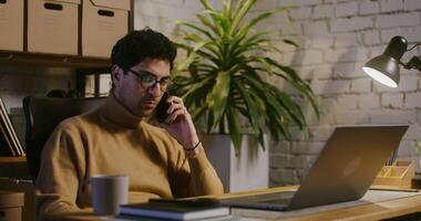 Young curly-haired dark-haired man wearing glasses with a serious look talking on a mobile phone, angry and cursing, sitting in front of an open laptop in a modern stylish office video