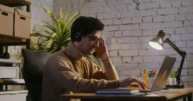 Young curly dark-haired man wearing glasses, with smiling talking on the phone using headphones with a microphone and typing text on a laptop while sitting at a table in the office video