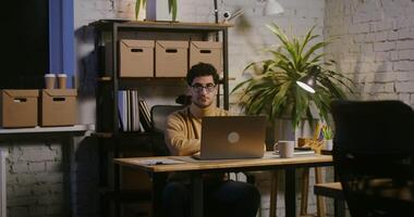 Young brunette man with stubble on his face looking straight into the camera and then taking off his glasses, while sitting at the table in front of an open laptop in a modern stylish office video
