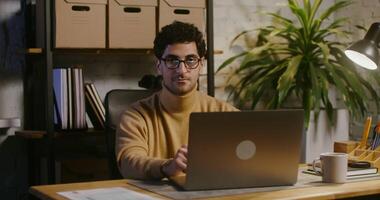 jovem morena homem dentro óculos com restolho em dele face, olhando em linha reta para dentro a Câmera enquanto sentado às a mesa dentro frente do a aberto computador portátil dentro uma moderno à moda escritório video