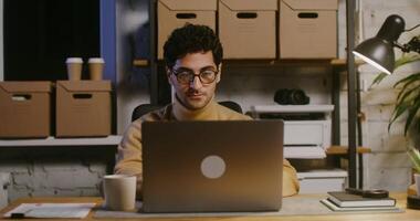Young handsome brunette man dressed in casual clothes takes off his glasses, while sitting in front of an open laptop and tired of work sitting at the table in a modern office video