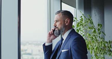 A man of European appearance, middle-aged, dressed in a business suit with a tie with a serious look, talks on a mobile phone, standing near the window in a modern office video