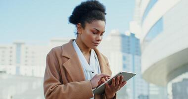 Beautiful young woman of African American appearance, dressed in a business style, is typing a message on the tablet and smiling looks around while standing outdoors in the business center of the city video