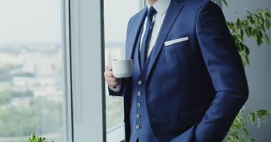 A man of European appearance with a beard, wearing a business suit with a tie, drinking coffee in the office and looking at his watch, close-up, the man's face is not visible video