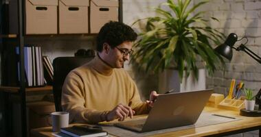 Young handsome curly haired brunette man, wearing glasses answers a mobile phone call and starts talking while sitting in front of an open laptop in a modern stylish office video