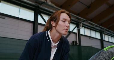 Young beautiful red-haired woman tennis player intently waiting for a pass from an opponent, ready to hit with a racket. Tennis match on the indoor tennis court video