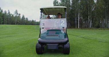 Two men of European appearance with a big age difference are driving a golf car on a golf course and talking to each other on a rainy cloudy day, front view video