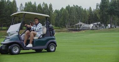 Two men of European appearance with a large age difference are driving a golf car across a golf course, talking to each other and looking around on a rainy cloudy day, side view video
