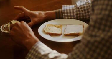 Young man of European appearance spreads butter on toast, preparing for breakfast, sitting at the table, close-up video