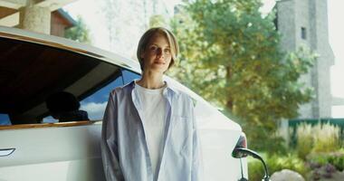 Beautiful young woman of European appearance, dressed in casual clothes, smiling while looking at the camera while standing near an electric car with a connected power cable near a modern mansion video