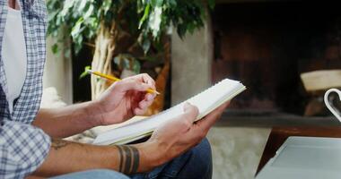 A man of European appearance, dressed in casual clothes, makes notes with a pencil in a notebook while sitting near an open laptop on a sofa in a modern interior, close-up video
