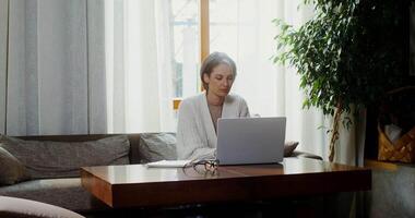 Beautiful young woman of European appearance, dressed in casual clothes, uses a laptop and takes notes in a notebook with her left hand, sitting in a modern home interior video