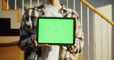A young woman of European appearance holds a tablet with a green screen in her hands, against the background of boxes while moving to a new home, close-up. Copy space, mock up video