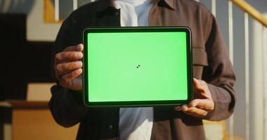 A man of European appearance holds a tablet with a green screen in his hands, against the background of boxes while moving to a new house, close-up. Copy space, mock up video