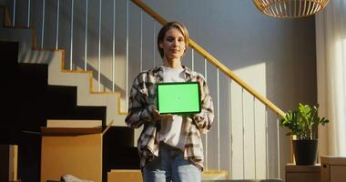 A young woman of European appearance holds a tablet with a green screen in her hands, against the background of boxes while moving to a new home. Copy space, mock up video