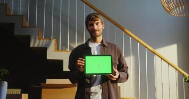 A man of European appearance smiles while looking at the camera and holding a tablet with a green screen in his hands, against the background of boxes while moving to a new home. Copy space, mock up video