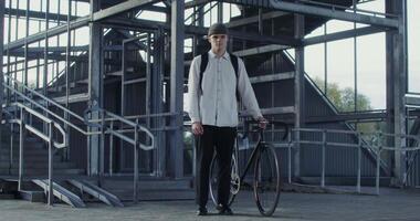 A young man of European appearance in a knitted hat and with a backpack on his back holds a bicycle with his hand and looks directly into the camera, standing near a metal ladder video