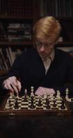 A young man of European appearance, dressed in a business suit, is studying with concentration to play chess alone, sitting at a table in the library, vertical shooting video