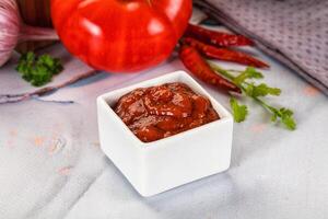 A small square container of tomato sauce next to a tomato and some herbs photo