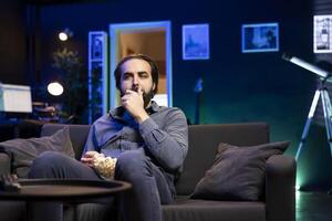 Lazy man enjoying leisure time at home watching TV, munching on popcorn. Couch potato in neon illuminated apartment, stuffing mouth with salty snack, entertained by television photo