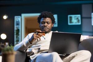 Man doing internet shopping while sitting on couch in living room, carefully typing credit card information on laptop. Person adding payment method on website while in RGB lit apartment photo