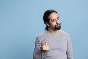 Rude irritated guy showing disrespect with the middle finger up, standing against blue background and acting like a hooligan. Male person with a negative impolite reaction does the offensive sign. photo