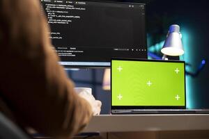 Man working from home using isolated screen laptop, testing and deploying programs and systems. Programmer checking code displayed on computer monitors using chroma key notebook photo