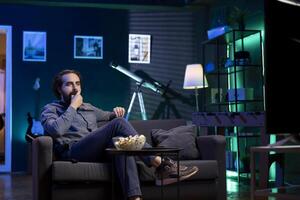 Cord cutter sitting on couch eating popcorn in front of TV displaying streamed movie. Upbeat person at home consuming snacks and watching on demand channels on television screen photo