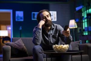 Cord cutter bored at home using TV to watch films on streaming services, zapping through content. Man in living room mindlessly consuming entertainment, using remote control photo