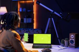 Woman in home office watching videos on social media using green screen laptop and phone. Person using social network website on chroma key notebook and smartphone, slacking off at work photo