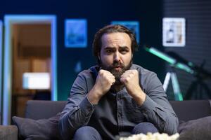 Man watching shows on TV, clenching punches, rooting for characters, paying close attention to screen. Man sitting on couch in apartment doing clasping hands gesture, hoping for happy ending photo