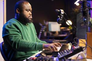 African american audio technician using piano synthesizer keys to add audio effects in post production, editing tunes with digital software and mixing console. Male artist in home studio. photo