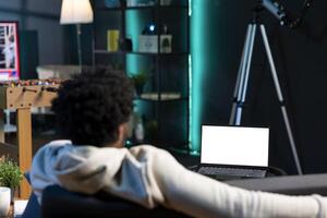 Man relaxing in living room by watching TV series on mockup laptop. BIPOC person enjoying leisure time in neon illuminated home, looking at multimedia content on isolated screen notebook photo