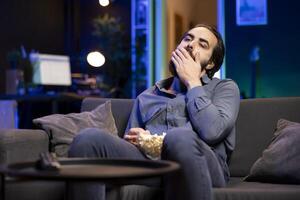 Bored man relaxing at home on couch, stuffing mouth with popcorn from bowl while watching movies on TV in apartment. Lazy person eating snacks and streaming shows in RGB lit living room photo