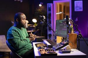 African american sound engineer using midi controller notes to add audio effects in post production, editing tracks with digital software and mixing console. Male musician in home studio. photo