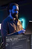 Joyful freelancer doing software debugging using laptop, checking errors in lines of code displayed on PC monitors. Cheerful IT professional walking in apartment fixing coding issues using notebook photo