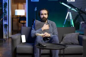 Frightened person on couch hugging pillow while binging suspenseful TV series on subscription based streaming services,. Afraid viewer at home watching horror movie on television display, eating snack photo