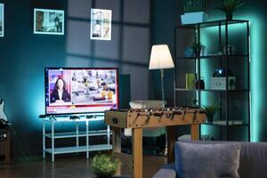 Dimly lit apartment with tabletop football in front of smart TV displaying news program broadcasting. Foosball table in living room and television screen in empty room showing media channel photo