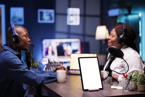 On desk, white screen with mockup template is displayed on tablet while African American vloggers are recording live podcast while wearing headsets. Digital device on table shows isolated chromakey. photo