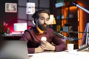 Content creator holding smart light bulb under face to create creepy mood in studio while telling horror story to audience. Entertainer in apartment using light to create atmospheric vibe photo