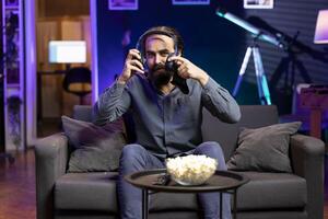 Smiling man putting headset on to play videogames in neon lit apartment, holding controller. Gamer participating in game using console system and headphones for immersive experience photo
