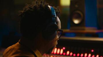 African american sound engineer using headphones to listen to audio recordings in post production, adjusting volume levels with editing software interface. Expert in studio control room. Camera A. photo