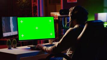 Man in dimly lit apartment playing games on green screen gaming PC at computer desk, enjoying day off from work. Gamer battling enemies in online multiplayer shooter on mockup monitor photo